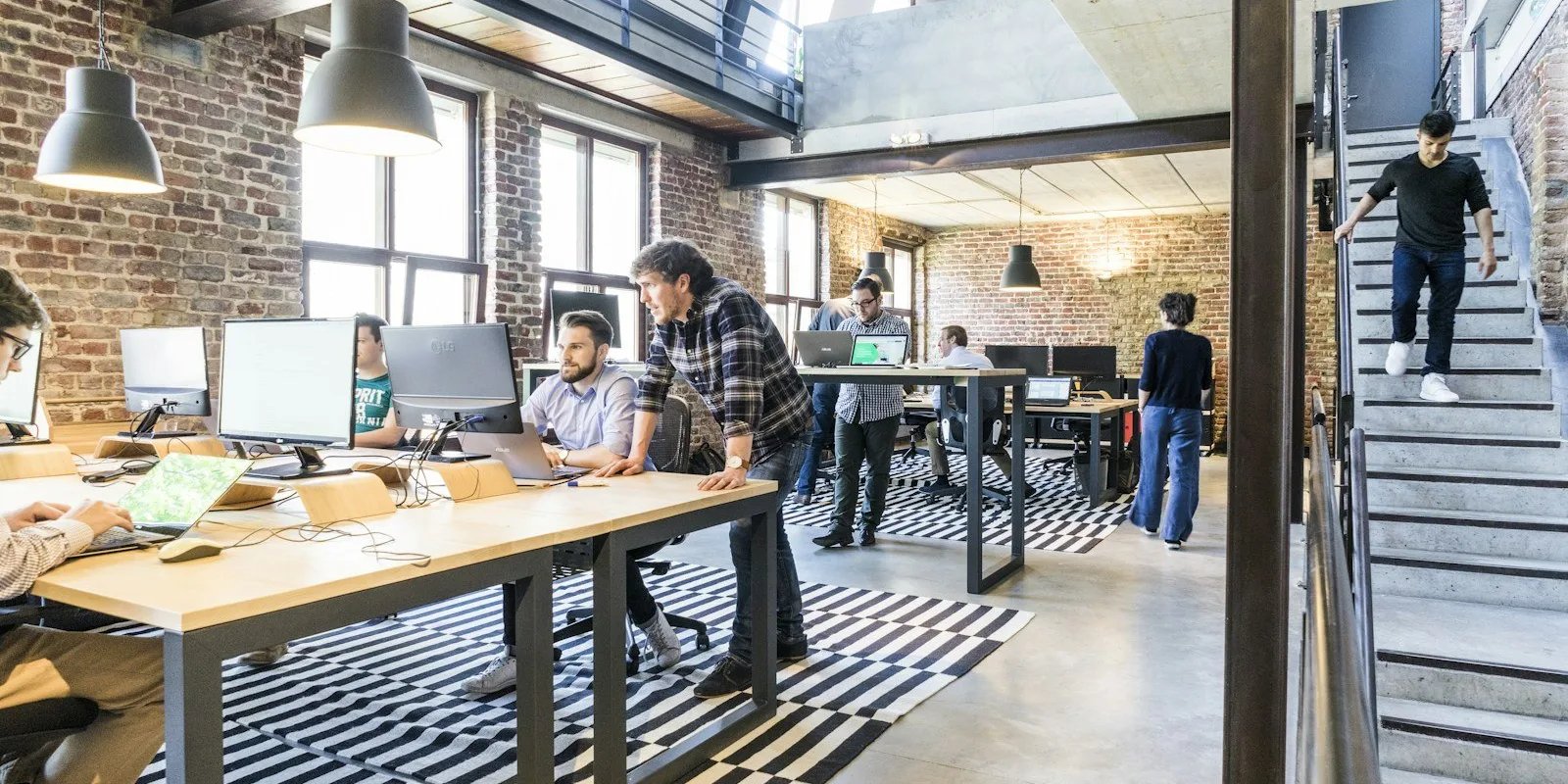 Offenes Büro eines Softwareteams: Ein Entwickler beugt sich über den Schreibtisch eines Kollegen, beide schauen konzentriert auf dessen Bildschirm